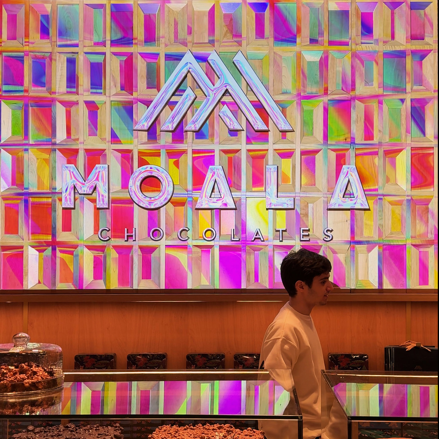 Logo Moala Chocolates proyectado en revestimiento de madera, paleta arcoíris