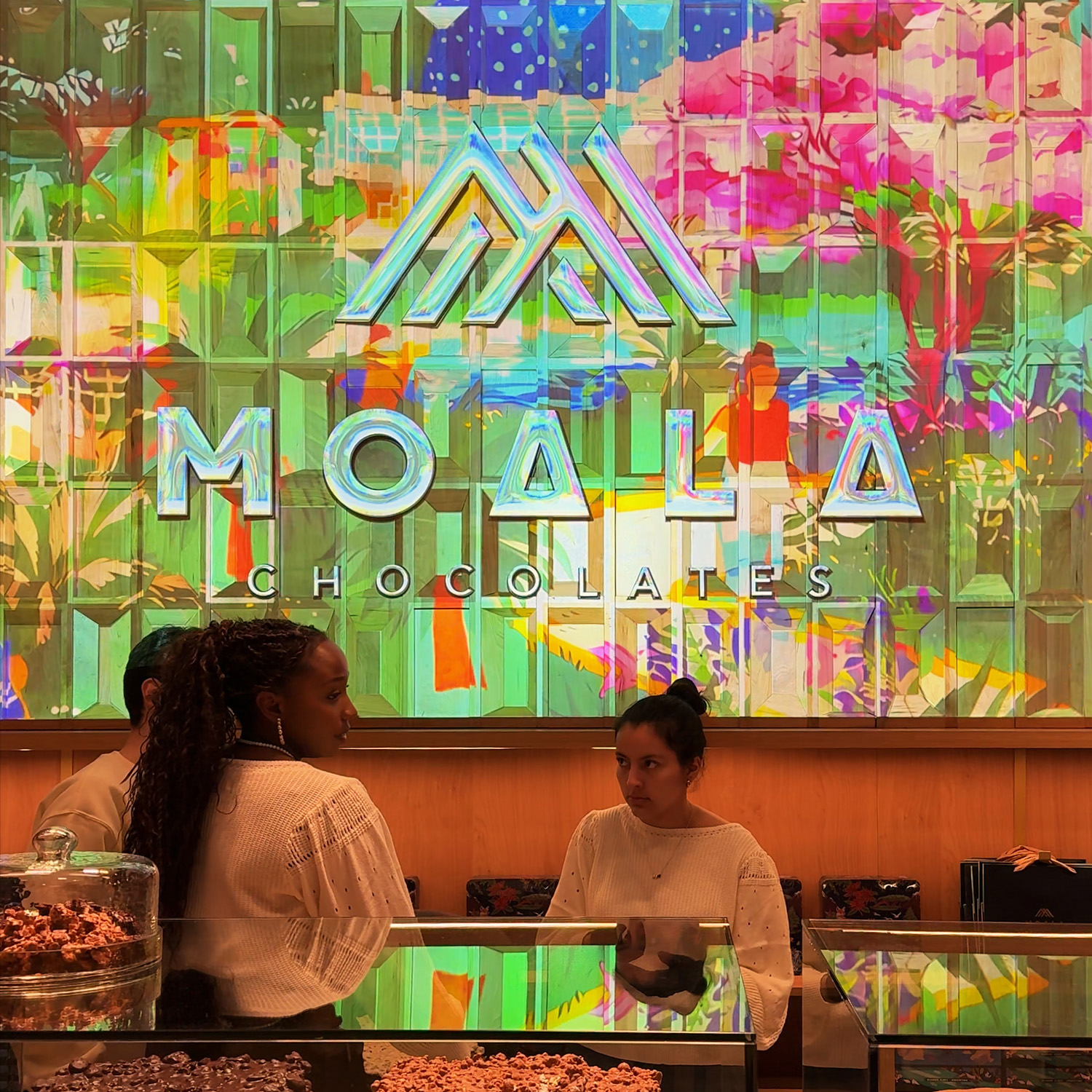 Logo Moala Chocolates proyectado en revestimiento de madera, paleta verde y flores